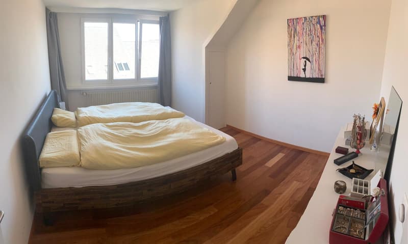 Bedroom with two beds, wooden floor, white walls, window with curtains, painting on the wall, desk with various items