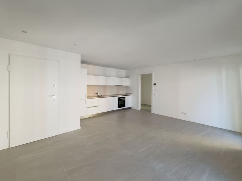 Empty room with white walls, tiled floor, kitchen area with sink, oven, and cabinets