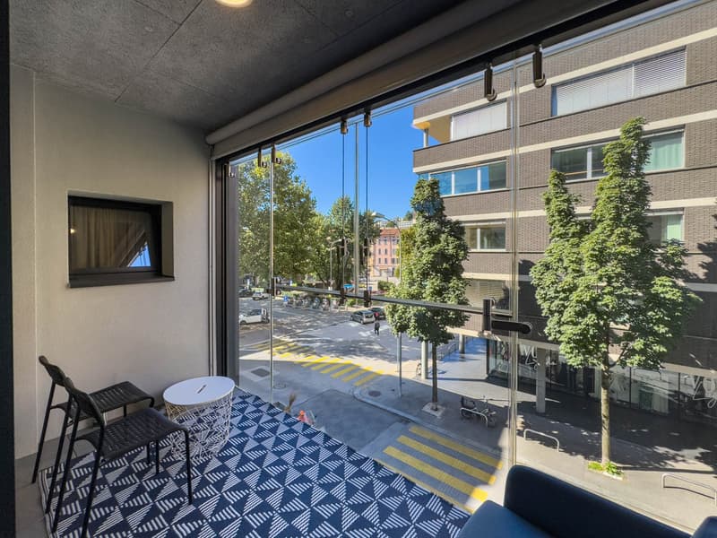 Balcony with glass railing, black chairs, white table, blue and white rug, city view