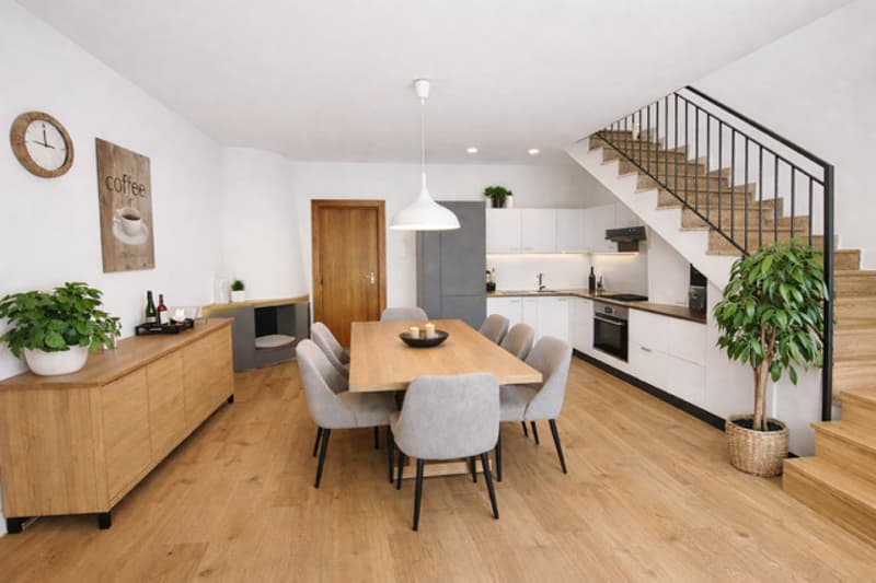 Dining room with wooden floor, dining table, chairs, wooden cabinet, kitchen with appliances, staircase, plants, and a clock on the wall.
