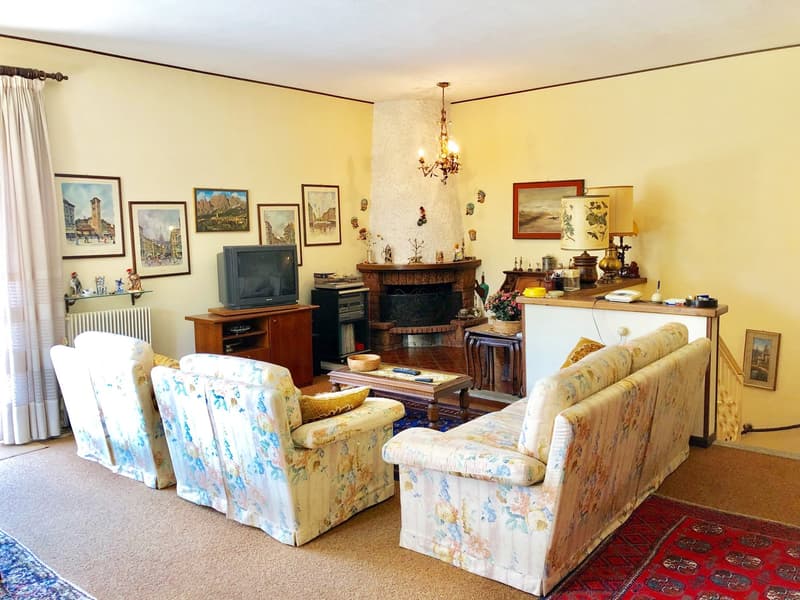 Cozy living room with floral patterned armchairs and sofa, wooden entertainment unit, fireplace, artwork on the walls, and a chandelier overhead