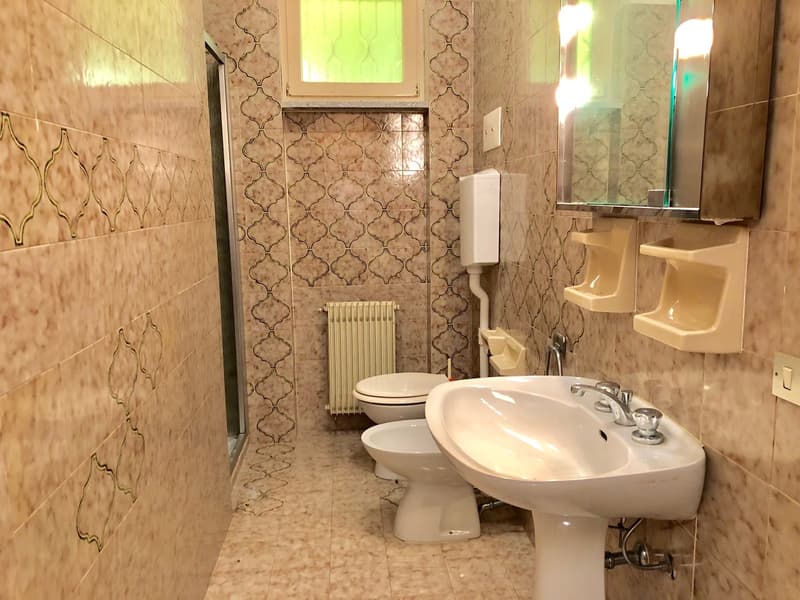 The image shows a bathroom with a tiled wall pattern featuring a quatrefoil design. The bathroom has a toilet, a pedestal sink, and a radiator. The room is well-lit with a window providing natural light.