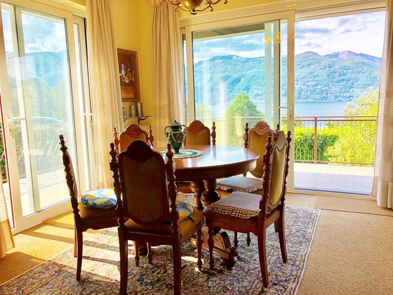 Elegant dining room with a round wooden table and ornate wooden chairs, overlooking a scenic mountain and lake view through large windows and a balcony. The room features a chandelier and decorative accents.