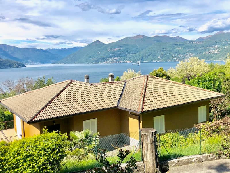 Single-story house with a tiled roof, surrounded by lush greenery and overlooking a scenic lake and mountains in the background. The house has a balcony or terrace offering a panoramic view of the beautiful natural landscape.