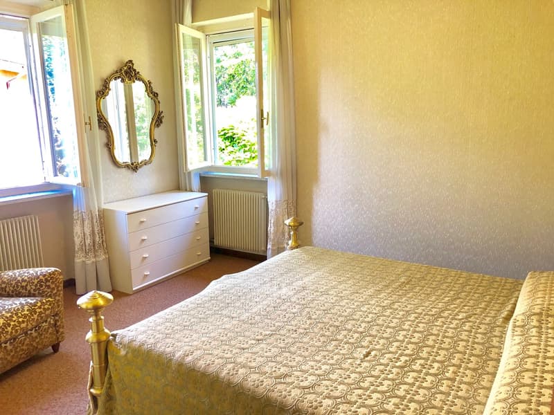 Spacious bedroom with large windows overlooking greenery, ornate gold-framed mirror, white dresser, patterned bedspread, and armchair.