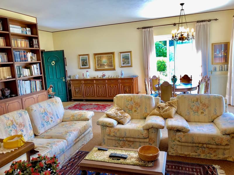 Spacious living room with floral patterned sofas and armchairs, wooden cabinet, bookshelves, framed artwork on the walls, and a chandelier overhead. The room has a cozy and traditional decor style.