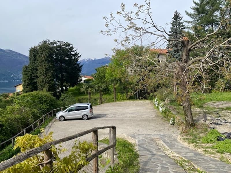 The image shows a scenic outdoor landscape with a paved walkway, trees, and a parked vehicle. The surroundings appear to be a rural or semi-rural area with mountains or hills in the background.