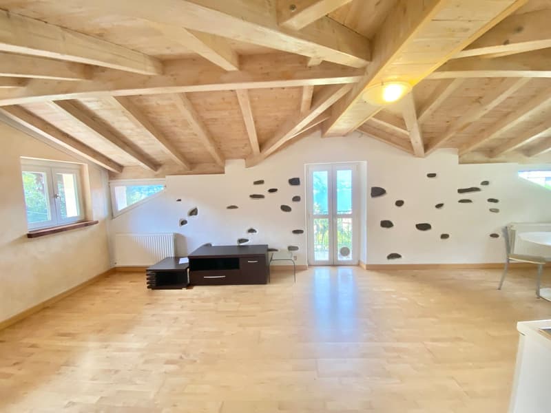 Spacious open-plan living room with exposed wooden beams on the ceiling, white walls, large windows, and a fireplace. The room has a minimalist design with a black entertainment unit and a few chairs.