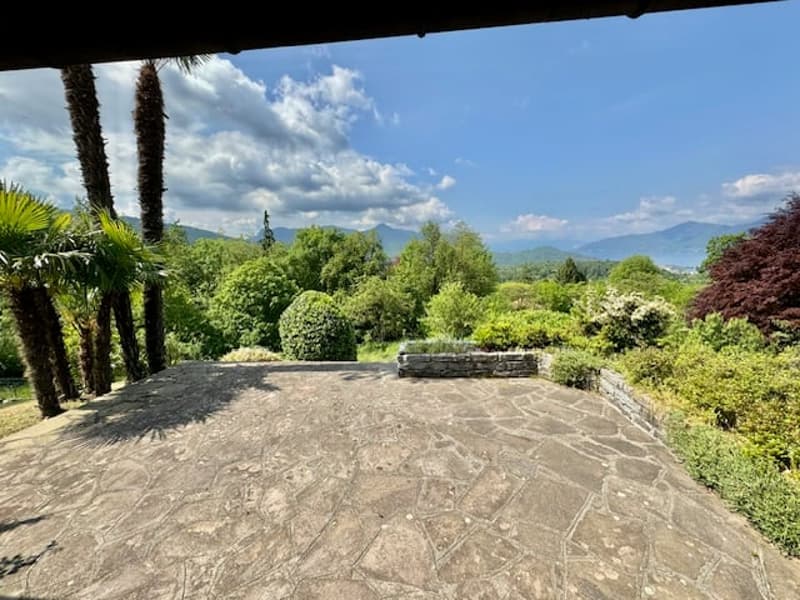 A spacious garden with a cobblestone path, surrounded by lush greenery, plants, and trees. In the distance, there are beautiful mountains under a cloudy sky.