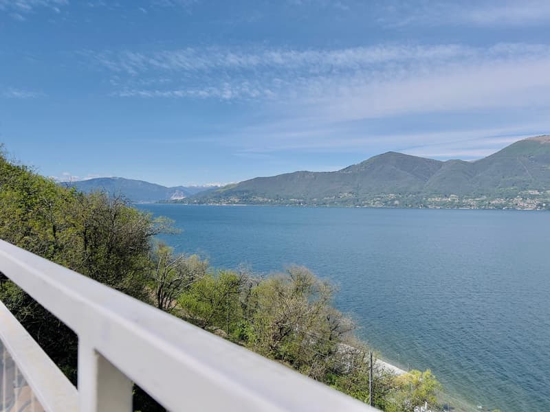 The image shows a scenic view of a lake surrounded by mountains. The lake appears to be a deep blue color, reflecting the clear sky above. The shoreline is lined with lush green vegetation, including trees and bushes. In the distance, the mountains rise u