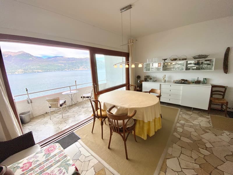Spacious dining room with large windows offering a scenic view of the lake and mountains. The room features a round dining table with wooden chairs, a sideboard, and shelving units displaying decorative items. The floor is covered with stone tiles, and th