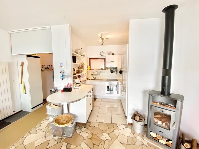 Open kitchen with white cabinets, stove, oven, and a small dining table. The floor is tiled with natural stone. There is a wood-burning stove in the corner of the room.