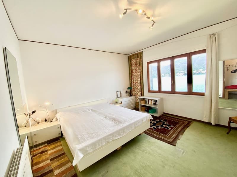 Spacious bedroom with large windows offering a scenic view, white walls, wooden furniture including a bed and shelving unit, and a patterned area rug on the floor.