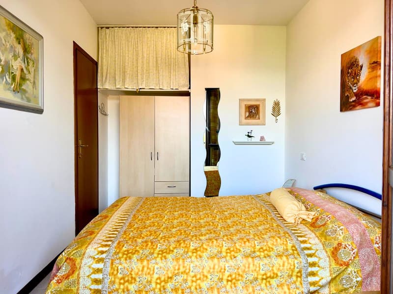 Spacious bedroom with a large floral-patterned bedspread, a wardrobe, and artwork on the walls. The room has a chandelier-style light fixture and a curtained window, suggesting the possibility of a balcony or terrace.