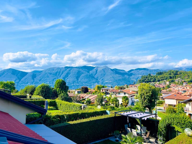 The image shows a scenic view of a town nestled among lush green hills and mountains. The town features a mix of residential and commercial buildings with red-tiled roofs, surrounded by well-manicured gardens and trees. The landscape is dominated by the t