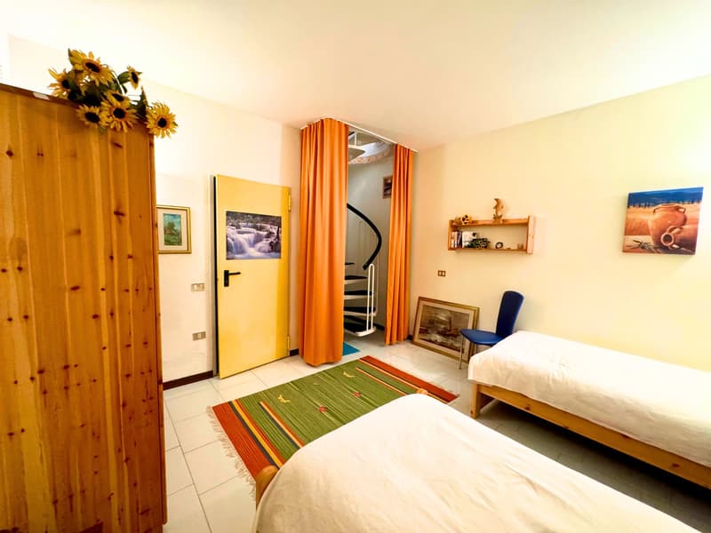 Cozy bedroom with wooden furniture, colorful rug, and sunflower decor. Large windows and orange curtains provide natural light. Artwork and shelving add personal touches to the space.