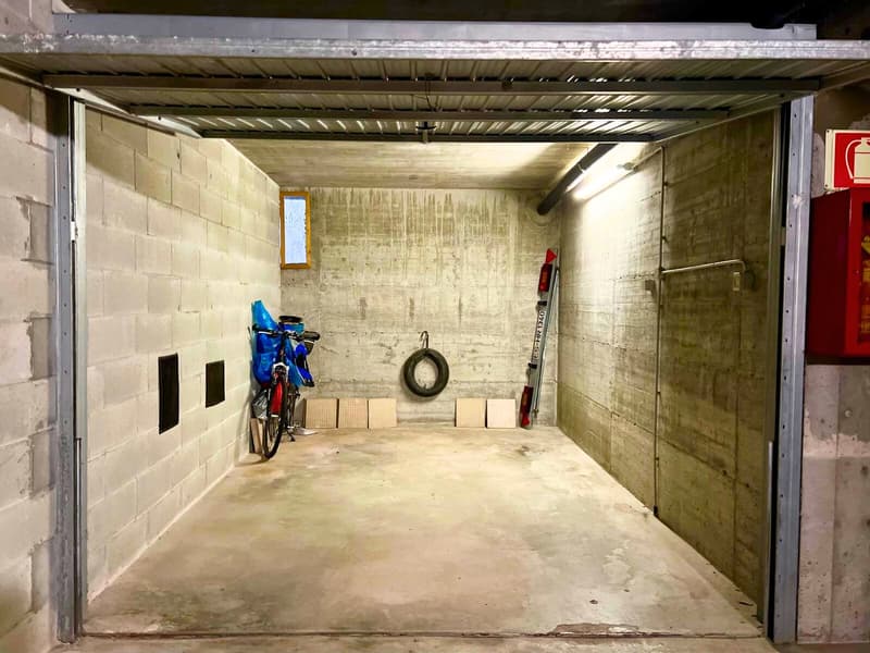 The image shows a garage or storage space with concrete walls and floor, a metal overhead door, and various items stored inside such as a bicycle, tires, and other equipment.
