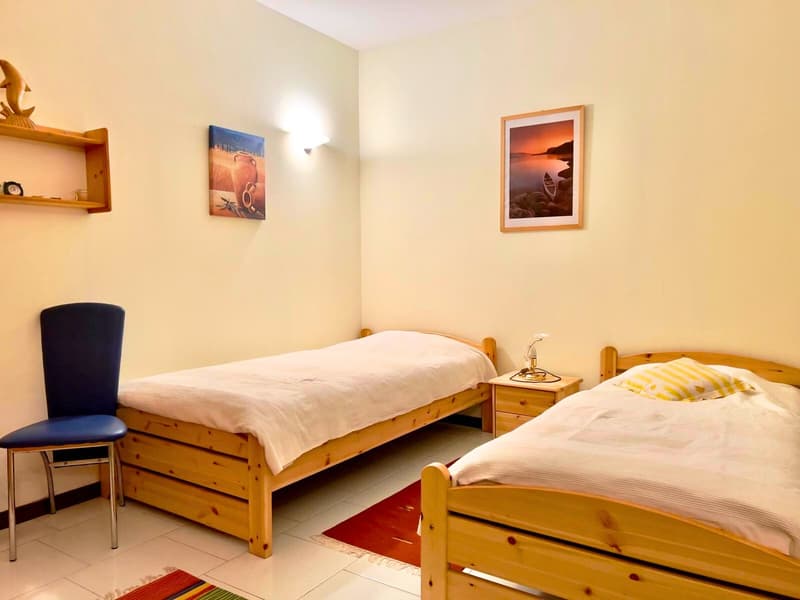 The image shows a bedroom with two twin beds, wooden furniture including a nightstand and chair, and wall art depicting scenic landscapes. The room has a warm, cozy atmosphere with neutral-colored walls and tile flooring.