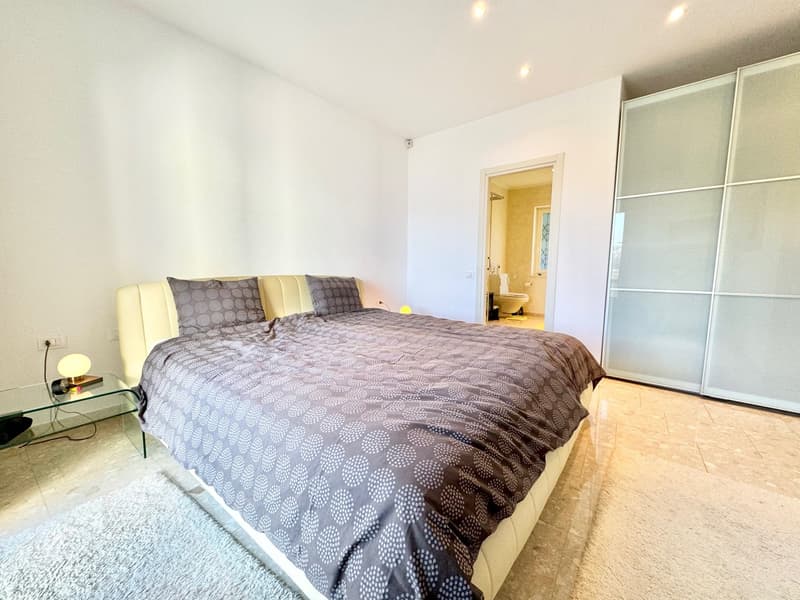 Spacious bedroom with a large bed, patterned bedding, and a sliding door leading to an ensuite bathroom. The room also features a glass-topped side table and a built-in wardrobe.