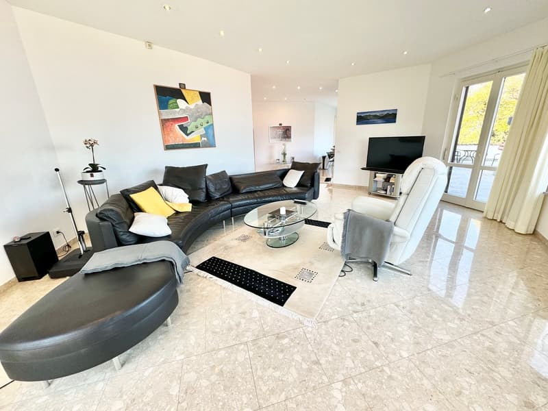 Spacious living room with large windows, curved leather sofa, glass coffee table, artwork on the walls, and a TV stand with a flat-screen TV.