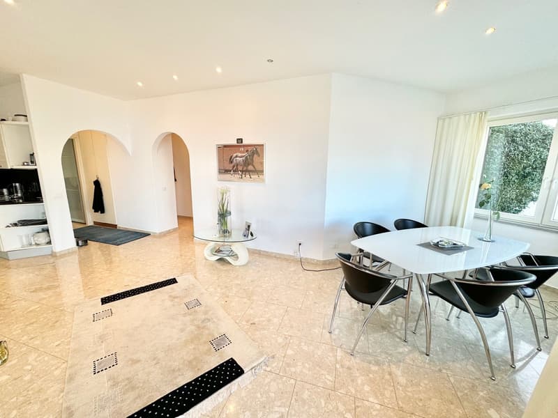 Spacious open-plan living area with white walls, arched doorways, tile flooring, a dining table with black chairs, and a decorative horse painting on the wall. The room has large windows that provide natural light and access to a balcony or terrace.