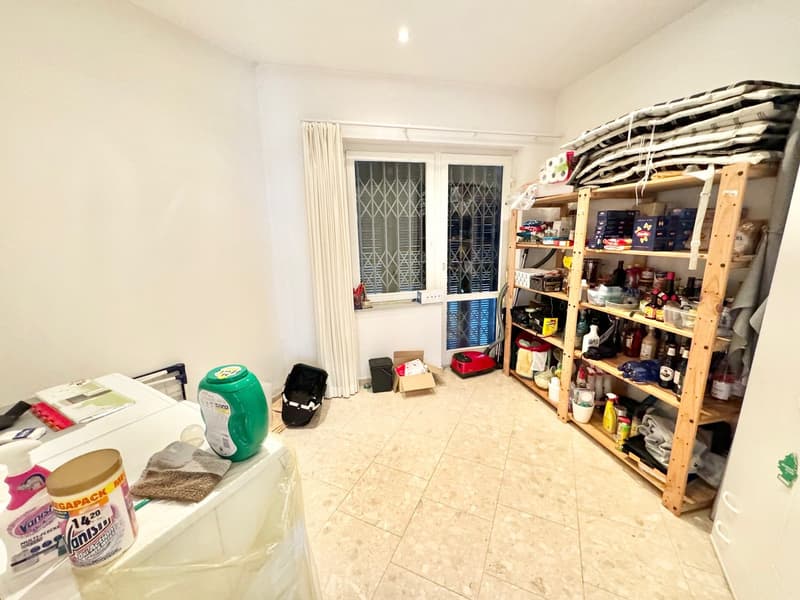 The image shows a storage room or utility room with various household items and supplies. There is a washing machine visible, as well as a shelving unit with various items stored on it. The room has tiled flooring and windows with decorative grilles.