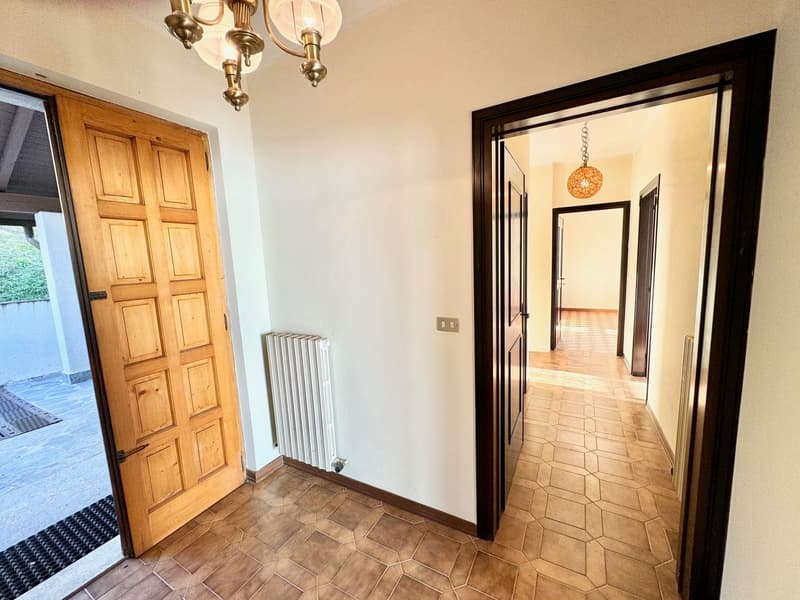 Hallway with brown floor tiles, white walls, a wooden door, open doors leading to different rooms, a white radiator, a chandelier, and two lamps.