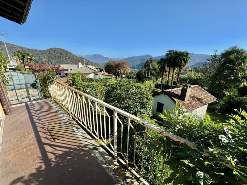 Two-story house with a balcony or terrace, surrounded by lush greenery and palm trees. The house has a tiled roof and is situated in a scenic mountainous area with a beautiful view.