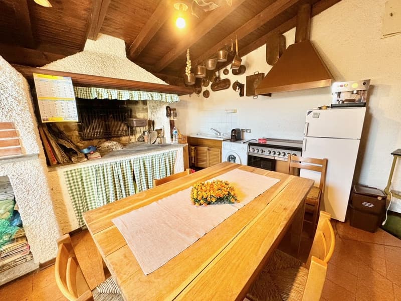 Rustic kitchen with wooden beams, a fireplace, a large wooden dining table, kitchen appliances including a stove and refrigerator, and various cooking utensils and decor hanging on the walls.
