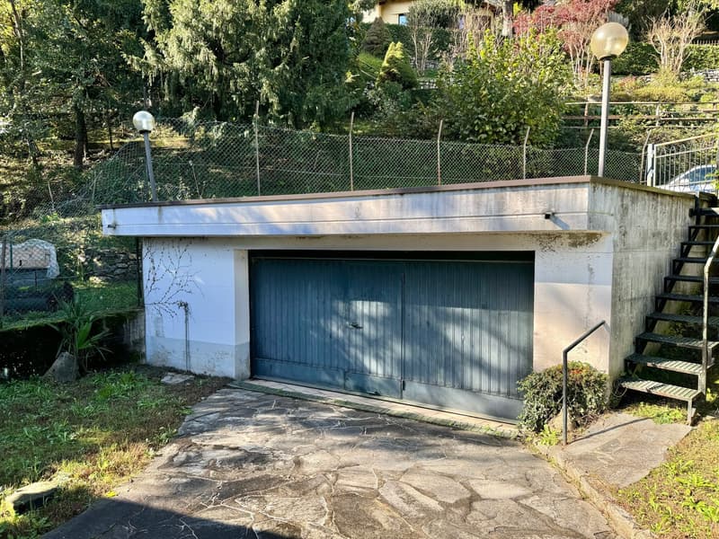 A concrete garage structure with a blue garage door, surrounded by lush greenery and trees.