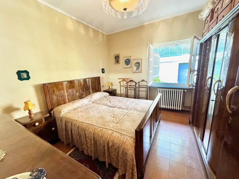 Spacious bedroom with a large wooden bed frame, bedside tables, and a chandelier overhead. The room has beige walls, a tiled floor, and a window that provides natural light. There are framed artwork on the walls and a small table with chairs near the wind
