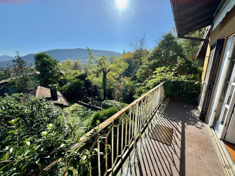 Wooden balcony overlooking a lush, green garden with trees and mountains in the background. The balcony has a wooden railing and appears to be part of a larger residential building or house.