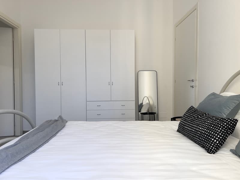 A bedroom with a large bed, white bedsheets, gray throw blanket, three decorative pillows, and a full wardrobe with drawers.