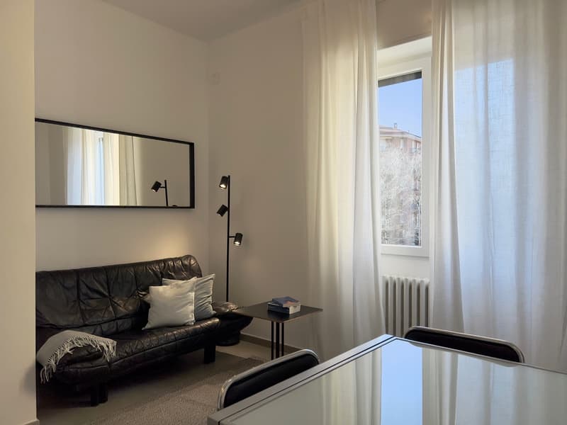 The image shows a living room with a black leather sofa, a side table, and a large mirror on the wall. The room has white walls and curtains, and a window that provides natural light. The lighting in the room is provided by a floor lamp with multiple spot