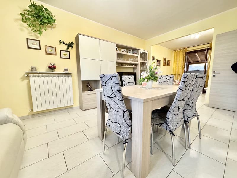 The image shows a kitchen and dining area. The kitchen features white cabinets, a tiled floor, and a radiator. The dining table has a light-colored wood top and patterned chairs. There are framed artwork and decorative items on the walls.