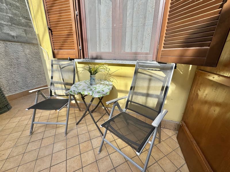 terraced room with tiled floor, 2 outdoor chairs, small table, plant, large window with wooden shutters, yellow wall
