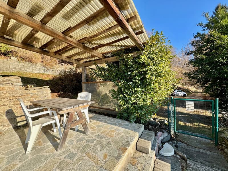 outdoor dining setup with a wooden table and chairs, stone-paved patio, covered pergola, stone wall, greenery, and a gate leading to a pathway with a parked car