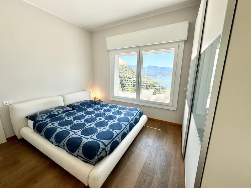 Spacious bedroom with large windows offering a scenic view of the mountains and lake. The room features a white leather bed frame, a patterned blue and white comforter, and hardwood flooring.