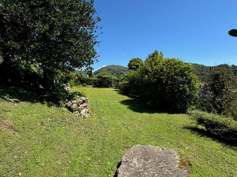 The image shows a lush, green garden with a well-maintained lawn. There are tall trees and shrubs surrounding the area, creating a serene and peaceful atmosphere. In the background, there appears to be a mountain or hill, suggesting a scenic view. The gar