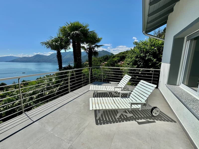 Spacious balcony with lounge chairs overlooking a scenic lake surrounded by mountains and lush greenery. The balcony has a modern, minimalist design with a glass railing and tiled flooring.