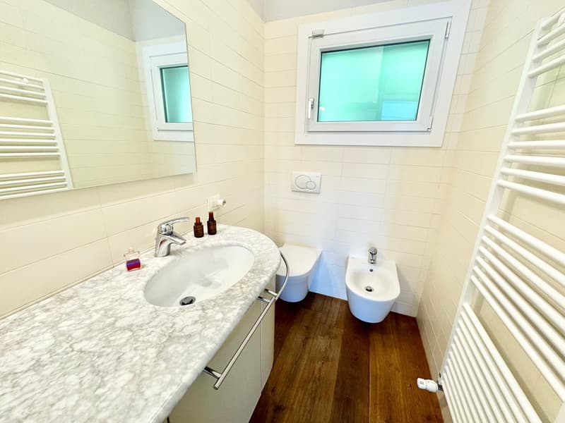 Bathroom with white tile walls, marble vanity countertop, sink, toilet, and a window with tinted glass. The floor is made of wood planks. There is a towel rack and a private washing machine visible in the room.