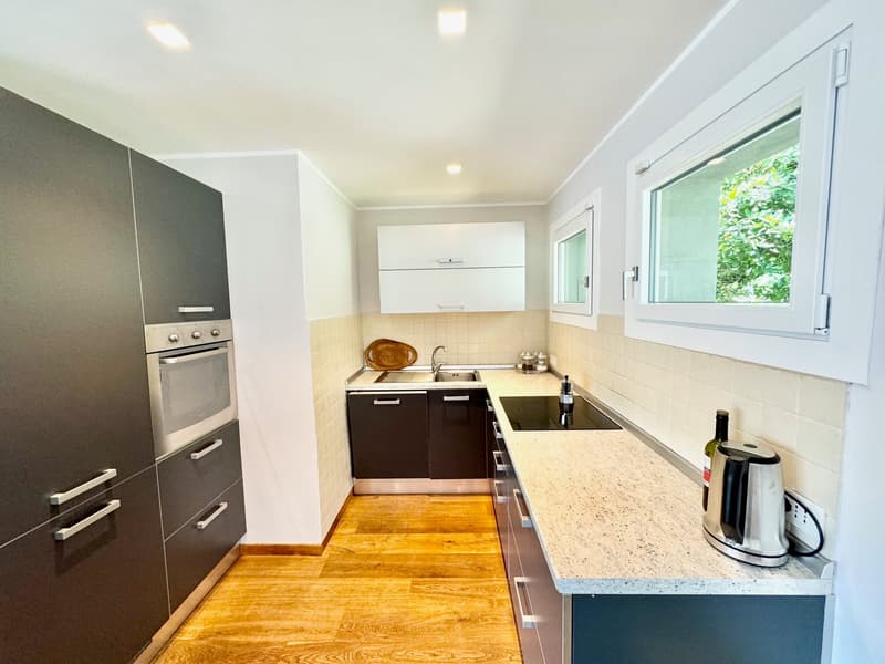 Modern kitchen with dark cabinets, white countertops, stainless steel appliances including a dishwasher, and a window overlooking greenery outside
