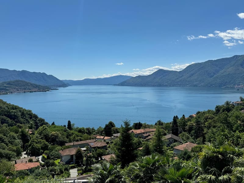 The image shows a scenic view of a lake surrounded by mountains and lush greenery. The lake appears to be a deep blue color, reflecting the clear sky above. The landscape is dotted with houses and buildings, nestled among the trees and vegetation. The ove