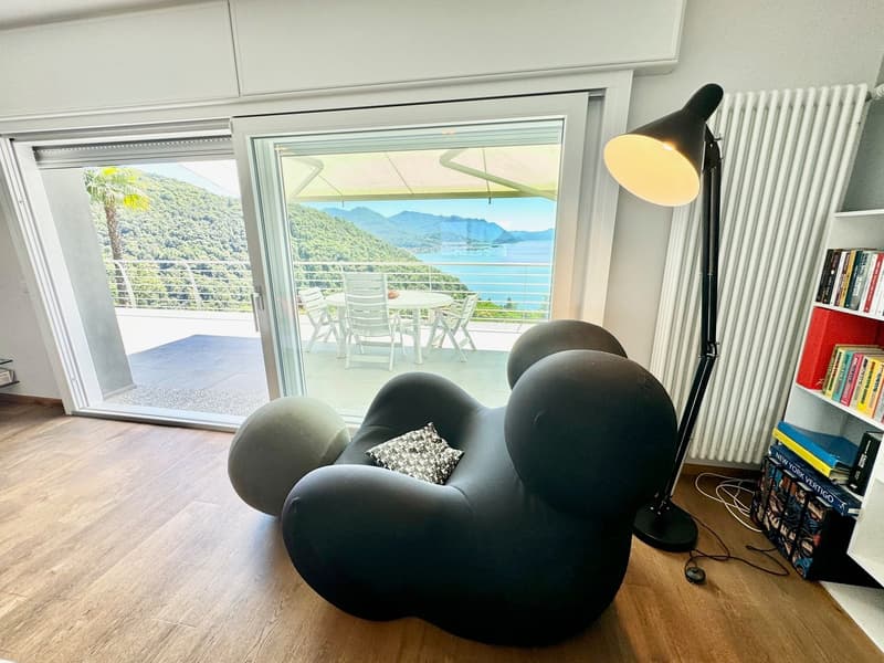 Spacious living room with large windows offering a scenic view of the mountains and lake. The room features comfortable black armchairs, a table, and a floor lamp. There is a balcony or terrace accessible through the sliding glass doors, providing an outd
