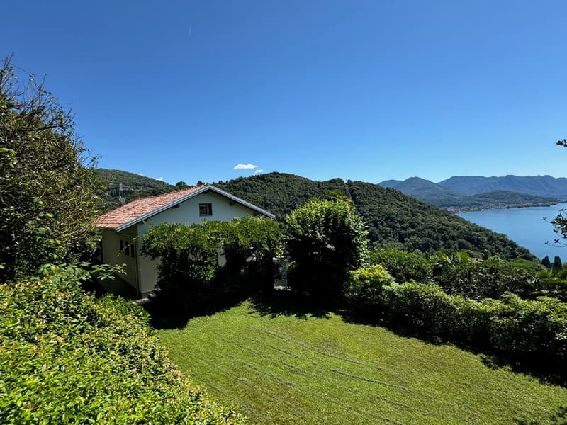Two-story house with a red tile roof, surrounded by lush greenery and vegetation. The house is situated on a hillside overlooking a lake or body of water, with mountains in the background. There appears to be a balcony or terrace on the upper level of the