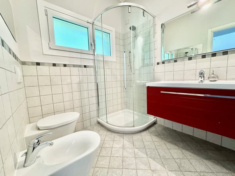 Spacious bathroom with tiled walls and floor, glass shower enclosure, vanity with red drawers, toilet, and window providing natural light