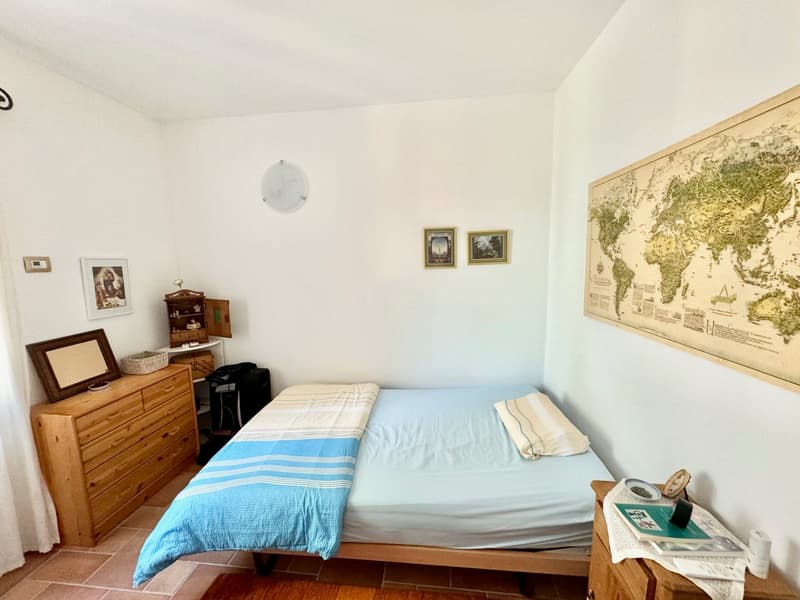The image shows a bedroom with a wooden dresser, a framed artwork on the wall, and a world map hanging on the wall. The room has a bed with a blue and white striped bedding, and there is a small table next to the bed with some items on it.