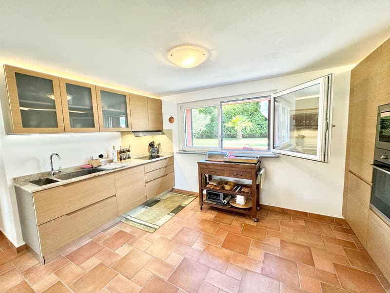 Spacious kitchen with wooden cabinets, large windows providing a view of the outdoor garden, tile flooring, and a movable kitchen island with shelves.