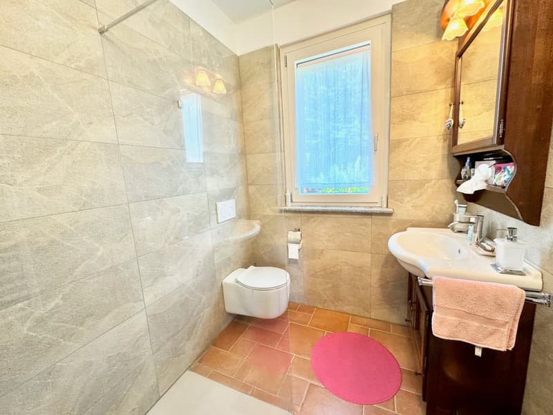 Spacious bathroom with large tiled walls, a toilet, a sink with vanity, and a window overlooking the outdoors. There is also a pink bath mat on the tiled floor.