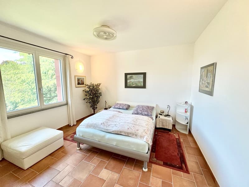 Spacious bedroom with large windows, hardwood floors, white walls, queen-size bed, nightstand, armchair, and artwork on the walls.